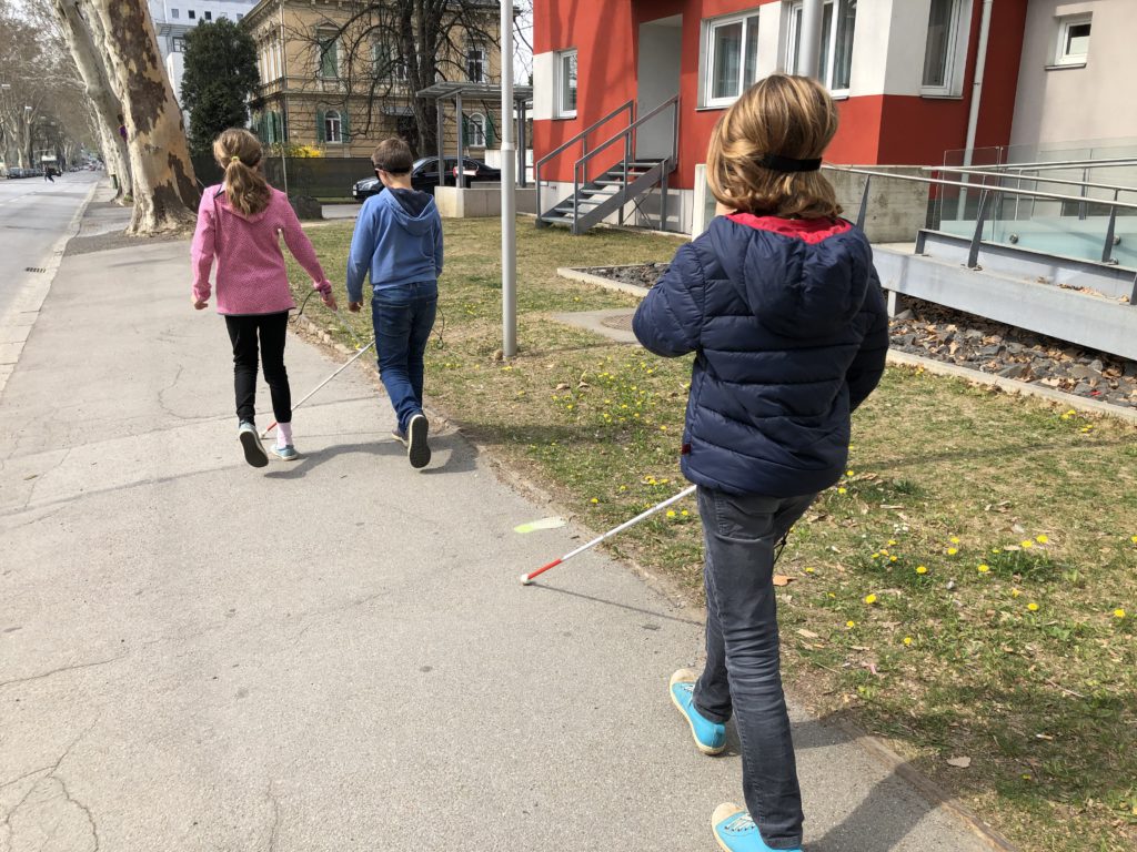 2 Schüler und eine Schülerin gehen mit Augenbinde und Blindenstock am Gehsteig.